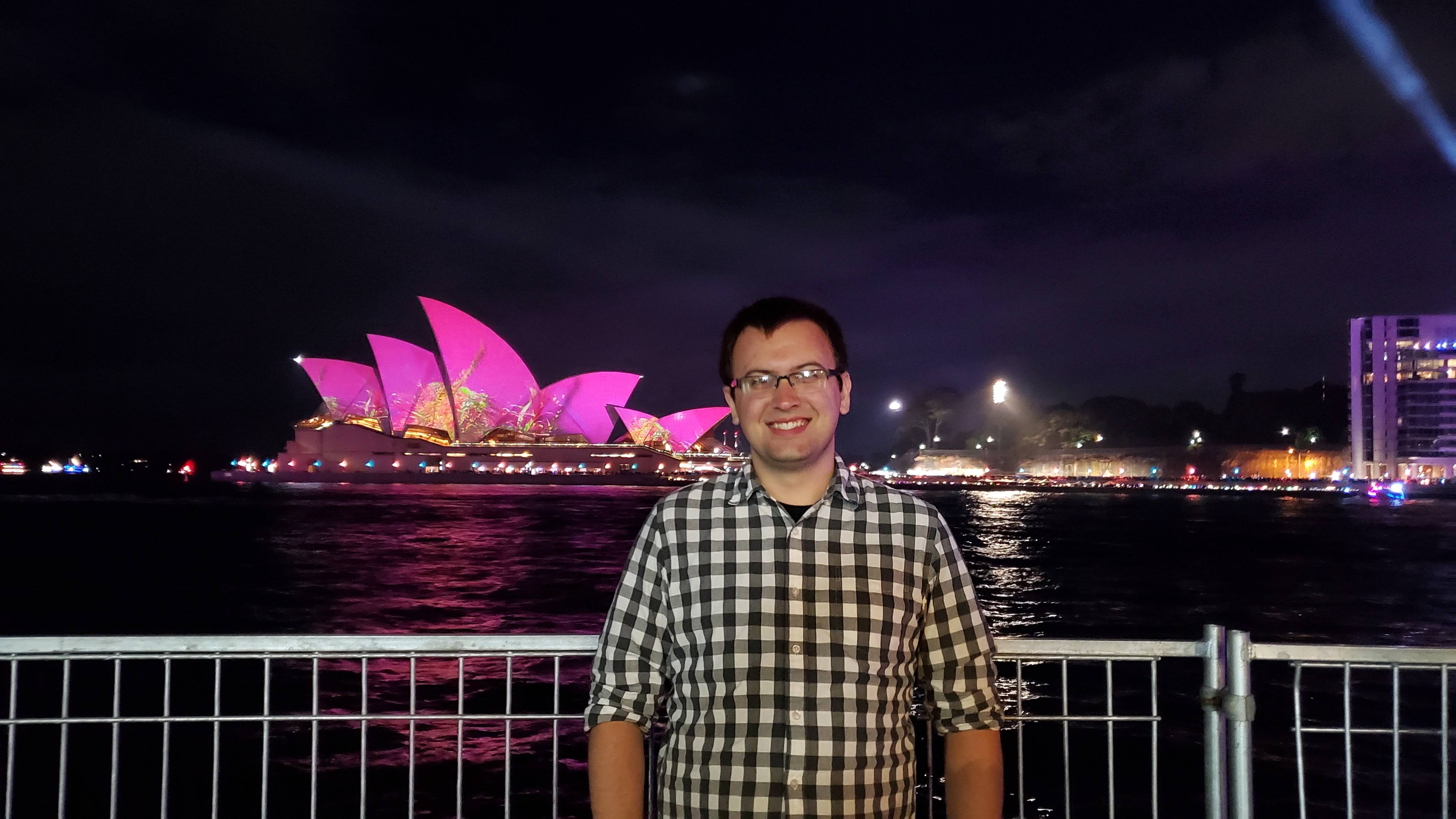 Kevin posing in front of Sydney Opera House illuminated in purple lights. 